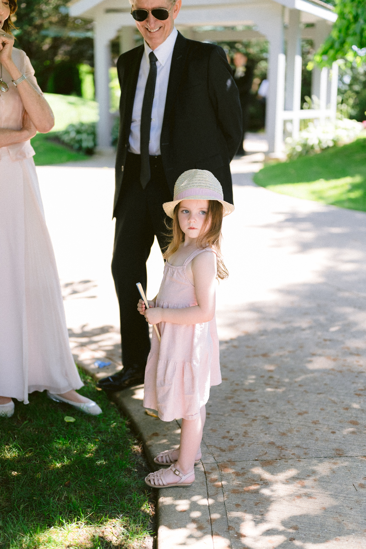 Young girl in a summer dress and straw hat standing in front of two adults at an outdoor gathering.
