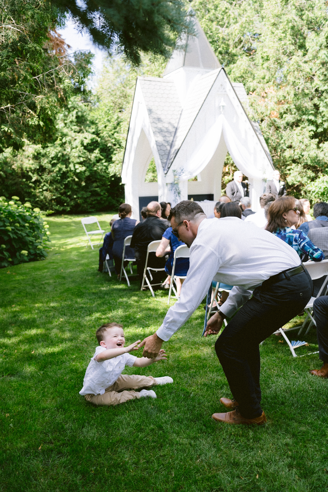 A man in a white shirt engaging with a small child on a grassy area at an outdoor event with guests seated facing a white canopy.