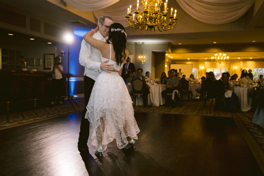 A couple dances together in an elegant ballroom with onlookers seated in the background.