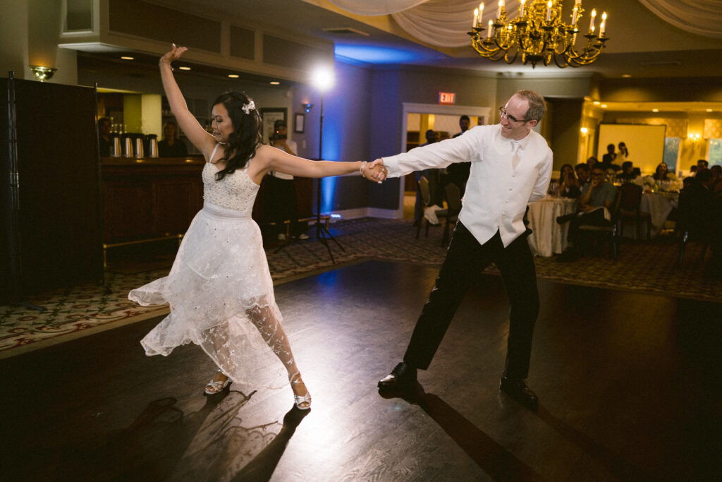 A couple dances together in an elegant ballroom with onlookers seated in the background.