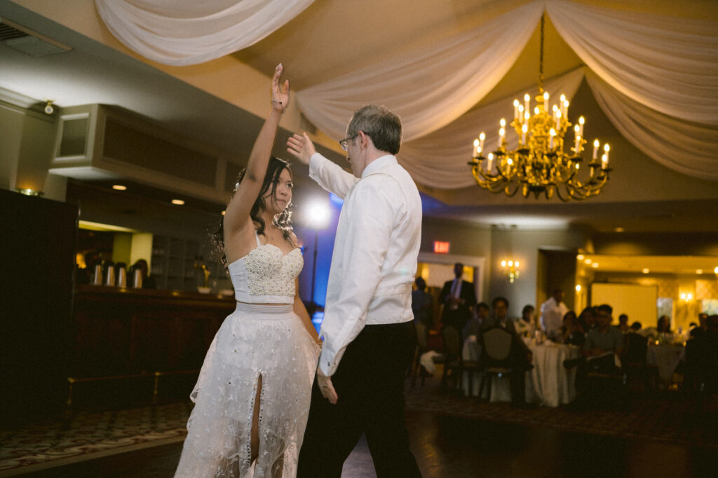 A couple dances together in an elegant ballroom with onlookers seated in the background.