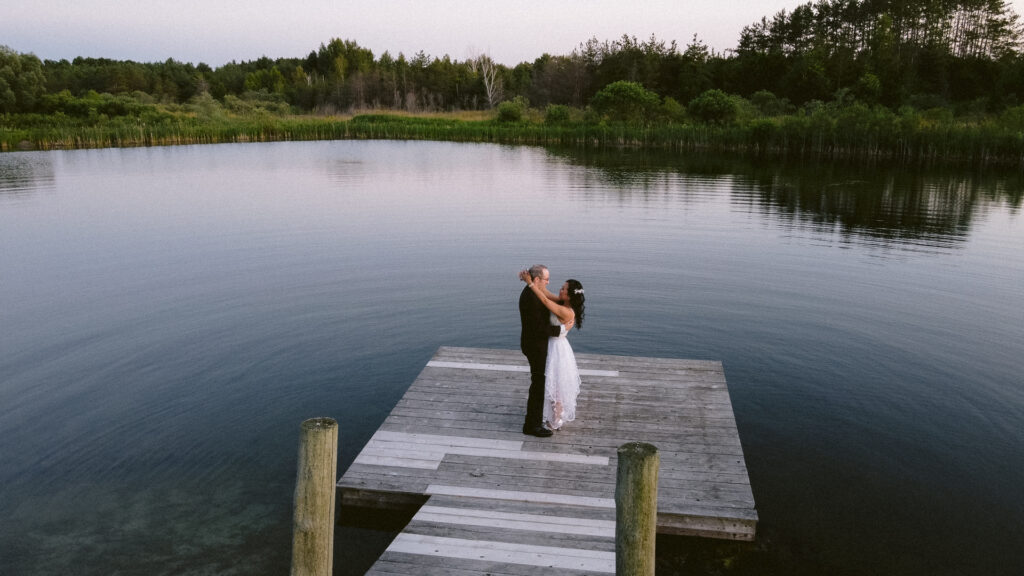 Couple embracing on a wooden dock by a calm lake with surrounding greenery at dusk