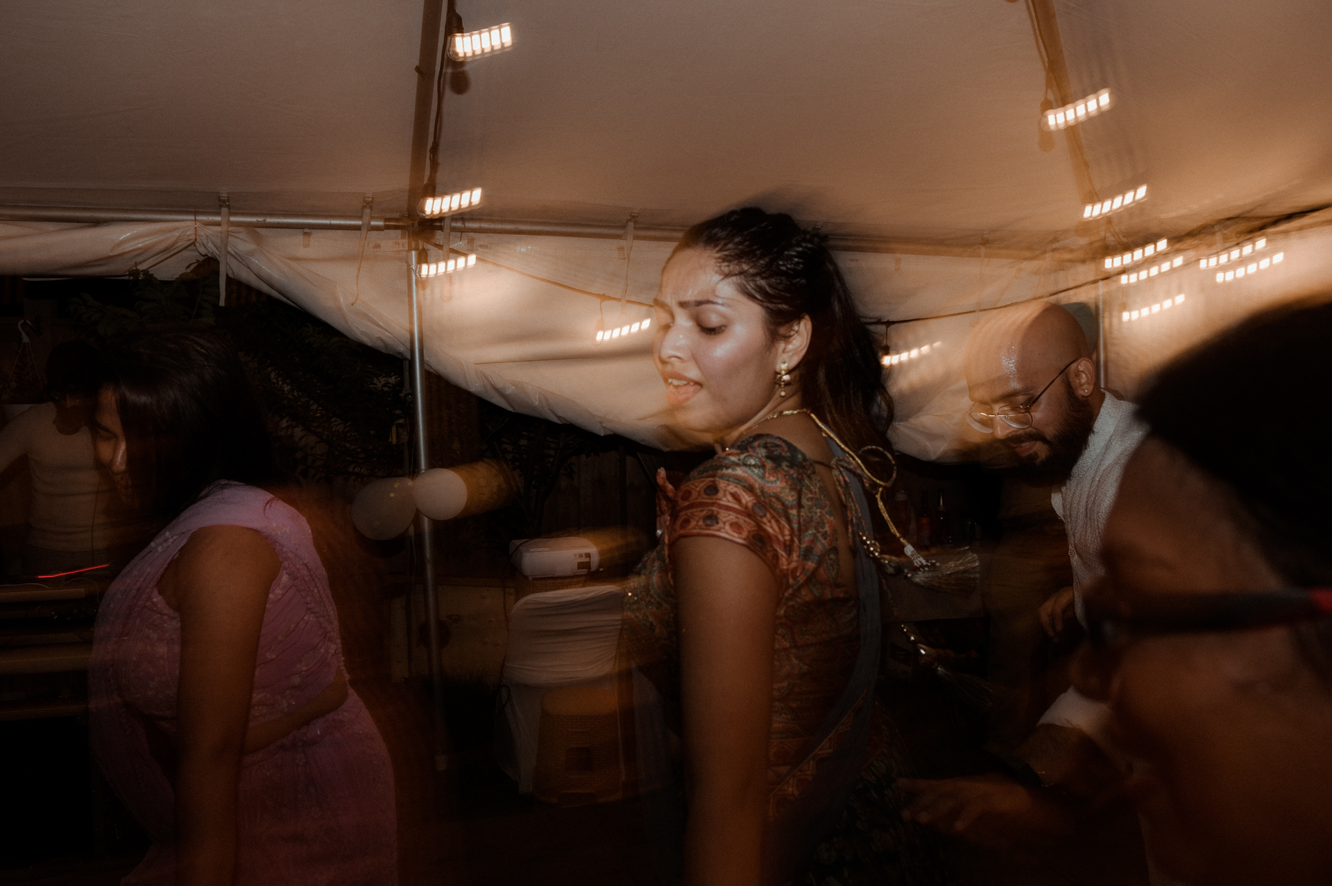 A blurred image of a woman dancing under a tent illuminated by string lights.