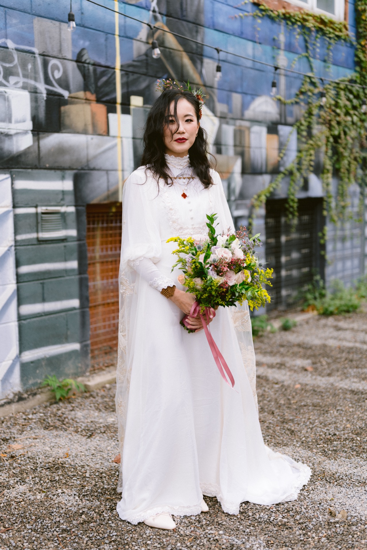A bride in a white dress holding a bouquet stands in front of a graffiti wall
