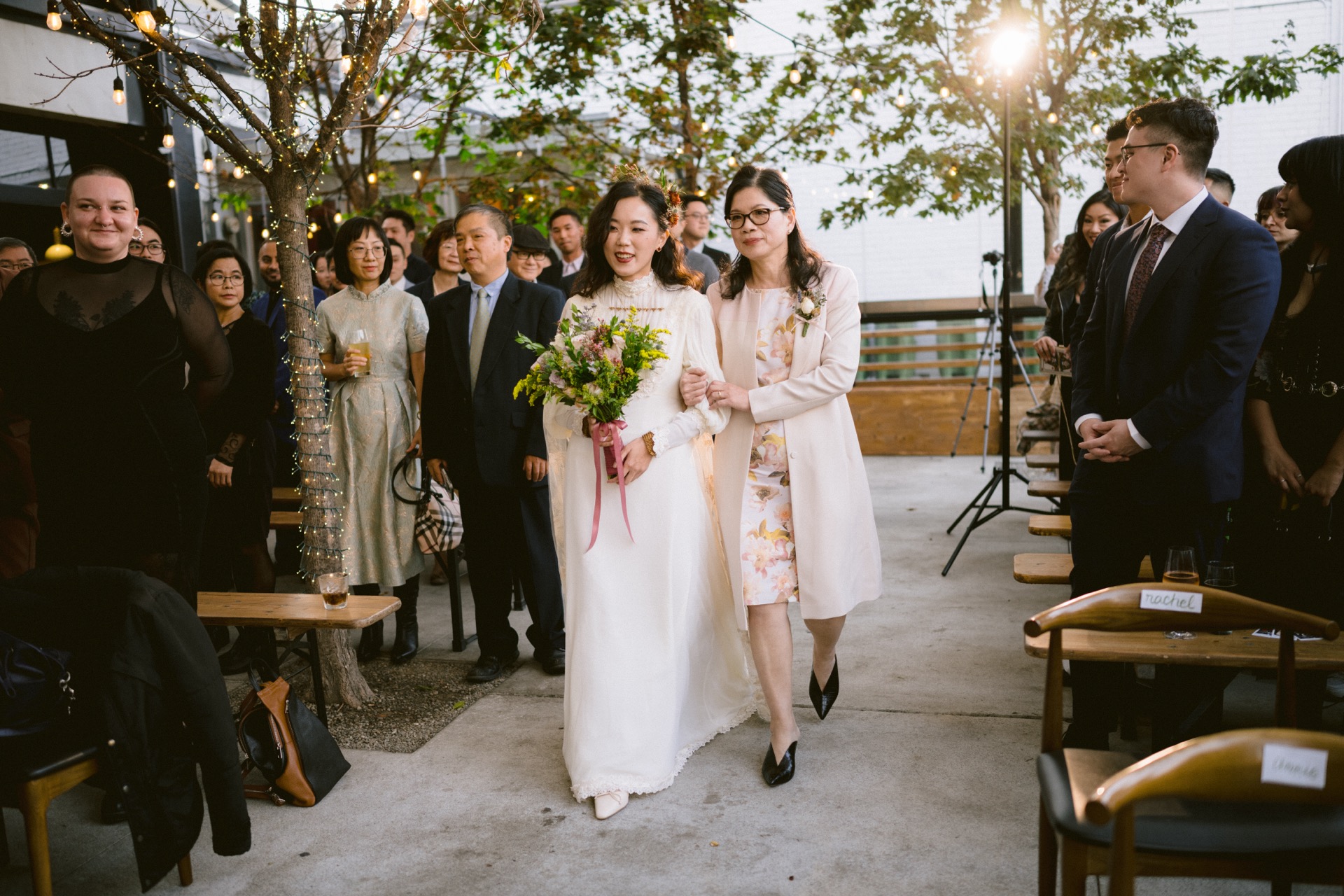 Two individuals in formal attire walking down an aisle at an outdoor wedding ceremony, with guests looking on and smiling.
