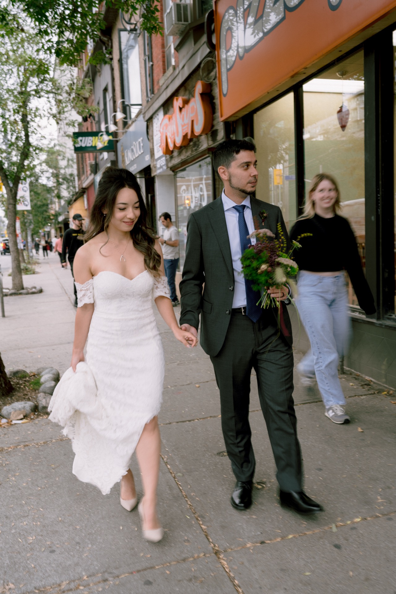 Couple in wedding attire holding hands while walking on a city sidewalk.