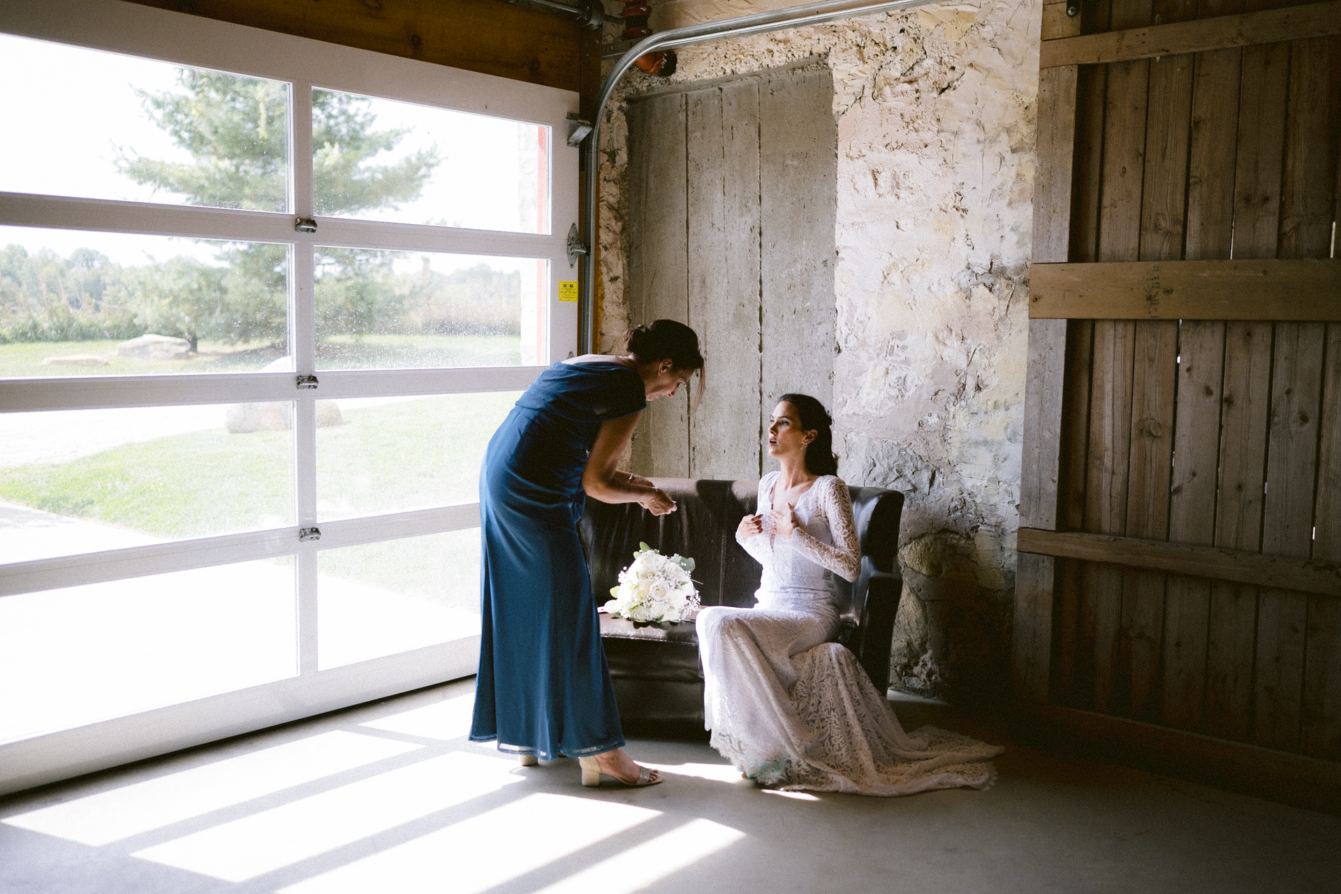 Bride's mother in a blue dress adjusts the veil of the bride seated in a chair, both are indoors by a large window, with a bouquet on the table next to them.