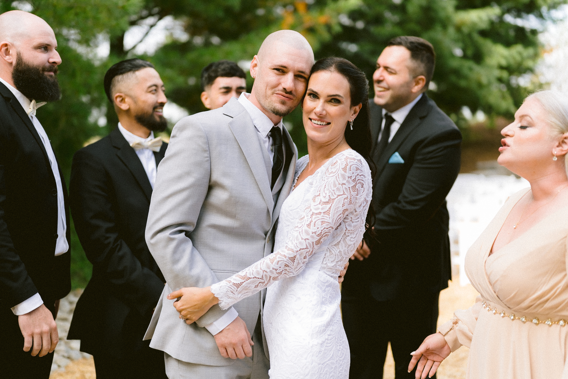 A couple in wedding attire embracing each other, with smiling guests in formal wear around them.