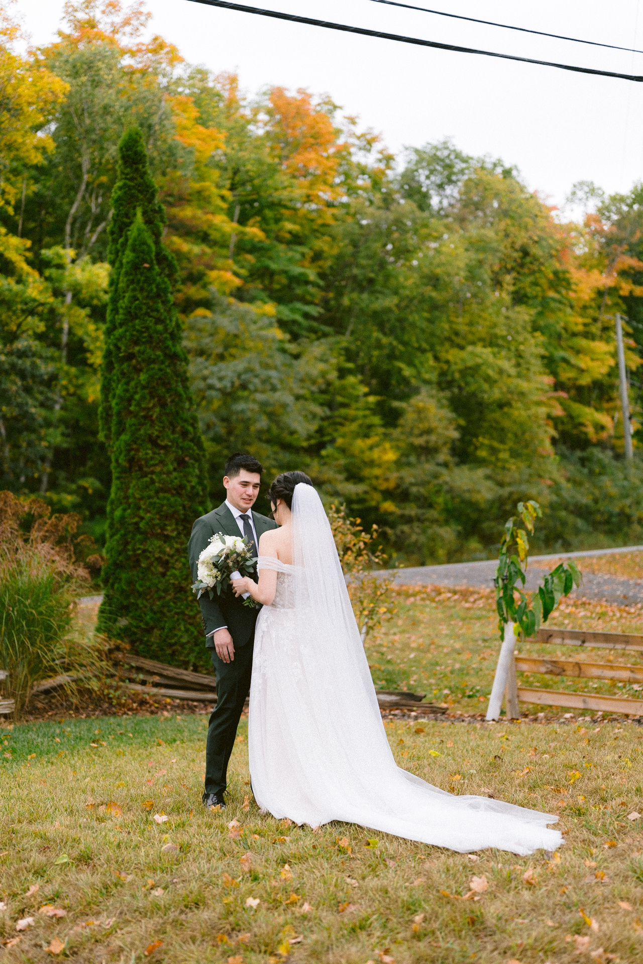 A bride and groom standing together on a lawn with autumn foliage in the background