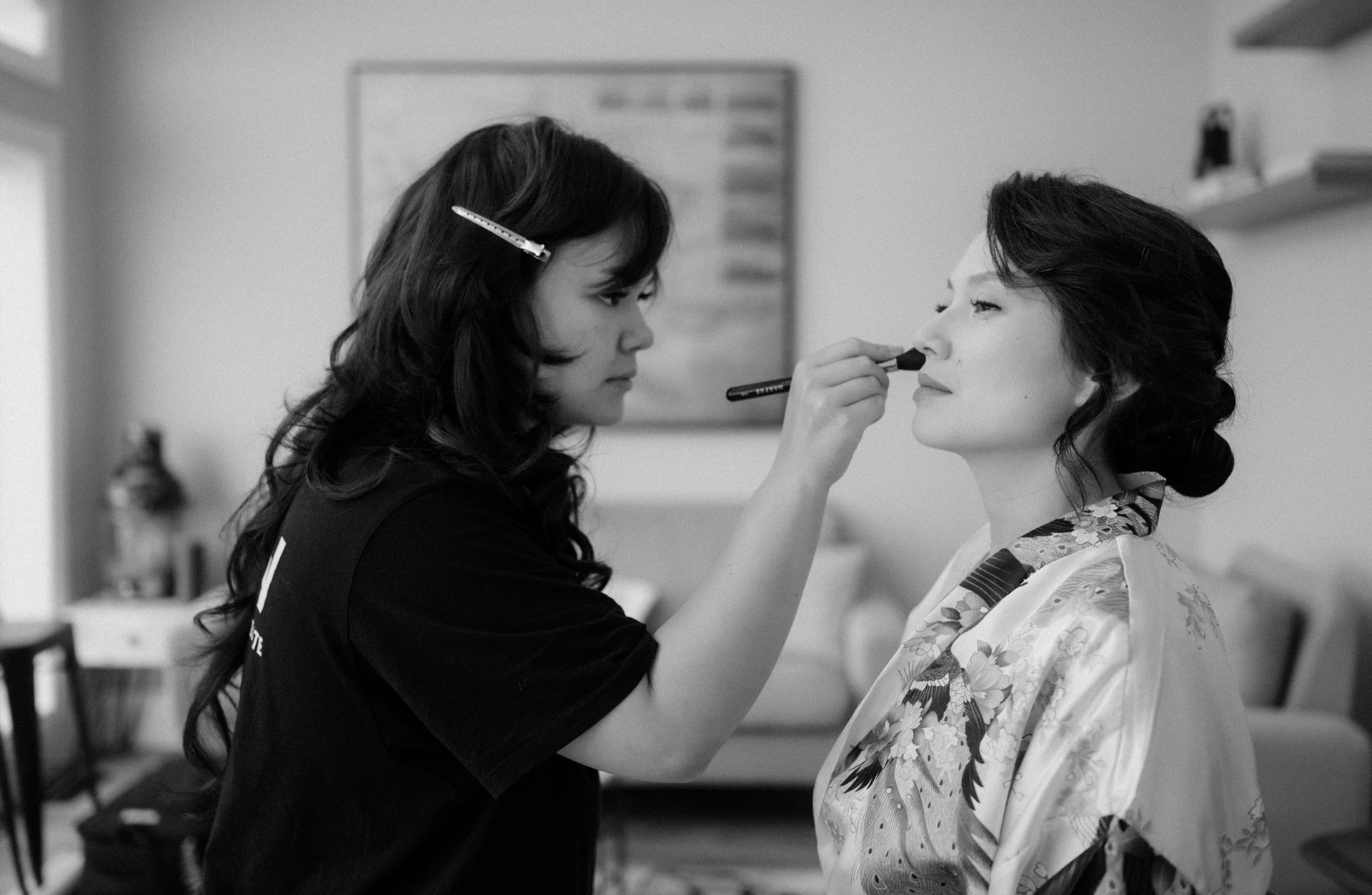 A makeup artist applying lipstick to a woman seated in a robe with a floral pattern for her fall wedding