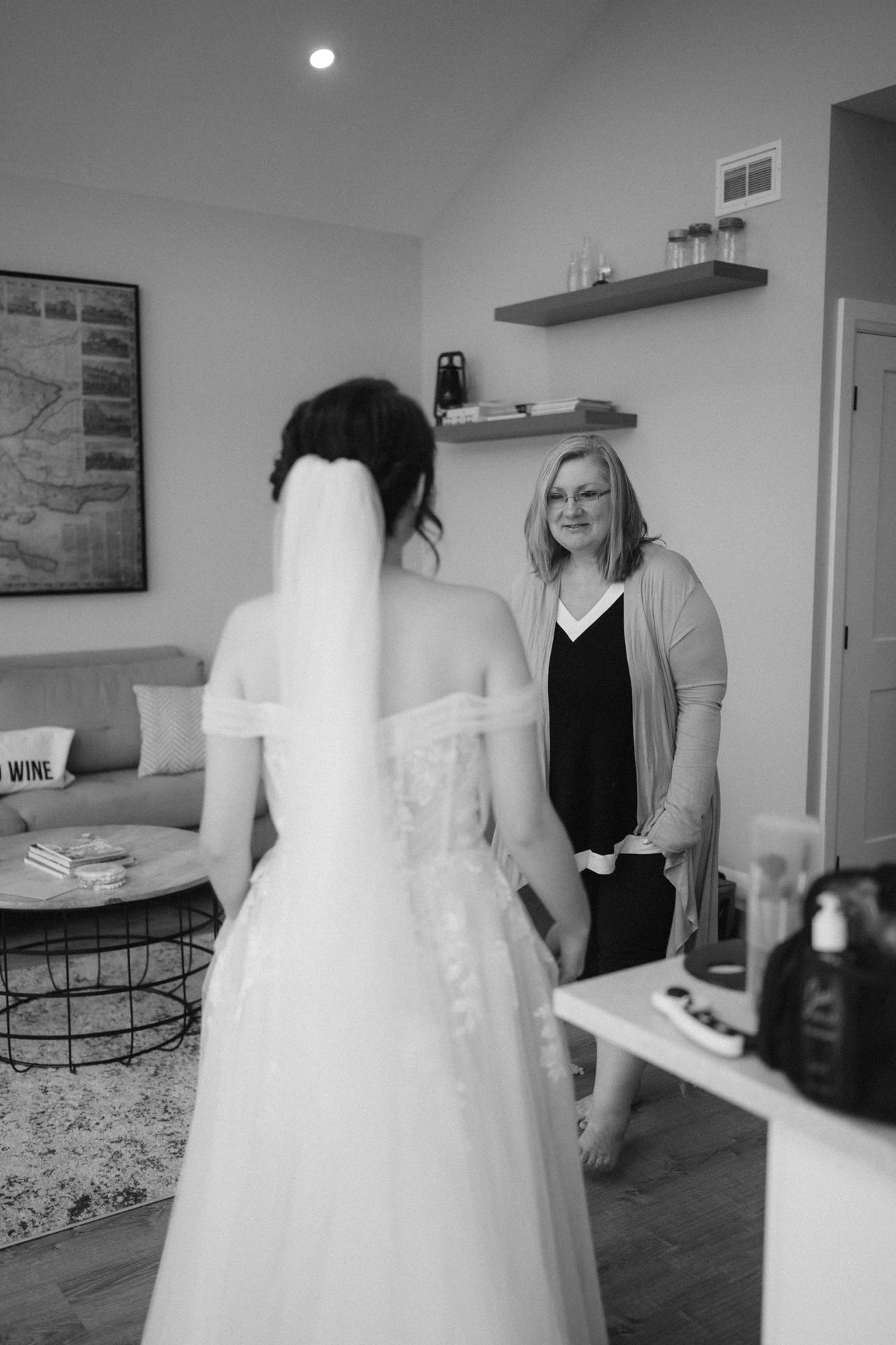 A bride in her wedding dress facing a smiling woman, likely a relative or friend, in a living room setting