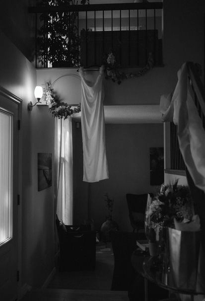 Monochrome interior of a room showing a staircase banister with a hanging weeding dress for a September wedding, framed photos on the wall, and a floral arrangement