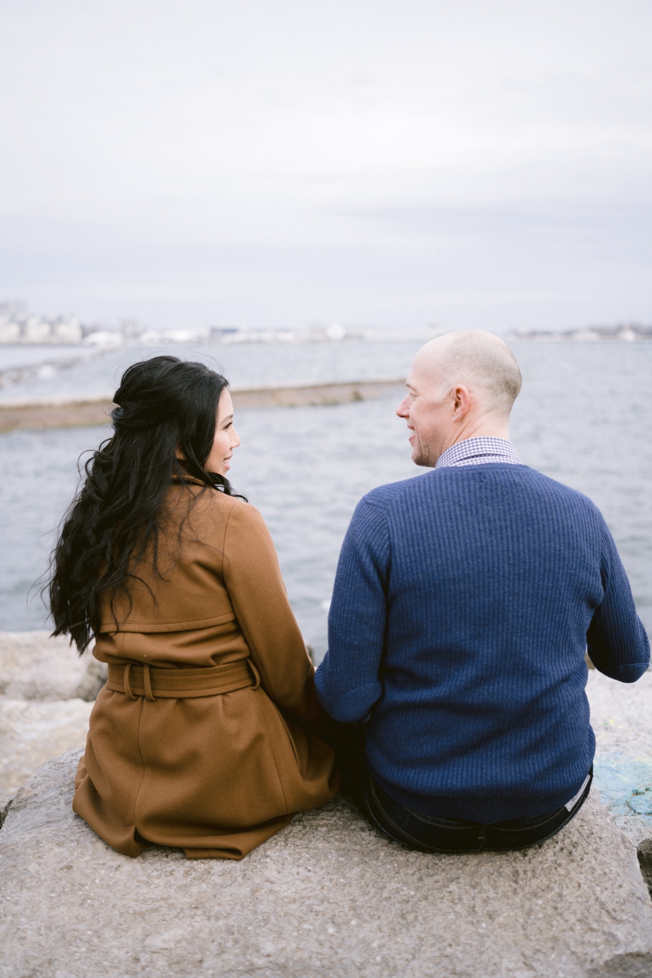 Two people sitting on a rock by the sea, looking at each other with a calm sea background