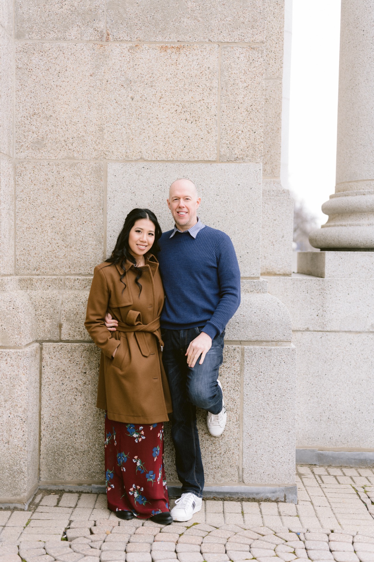 A smiling man in a blue sweater and jeans standing by a stone wall.