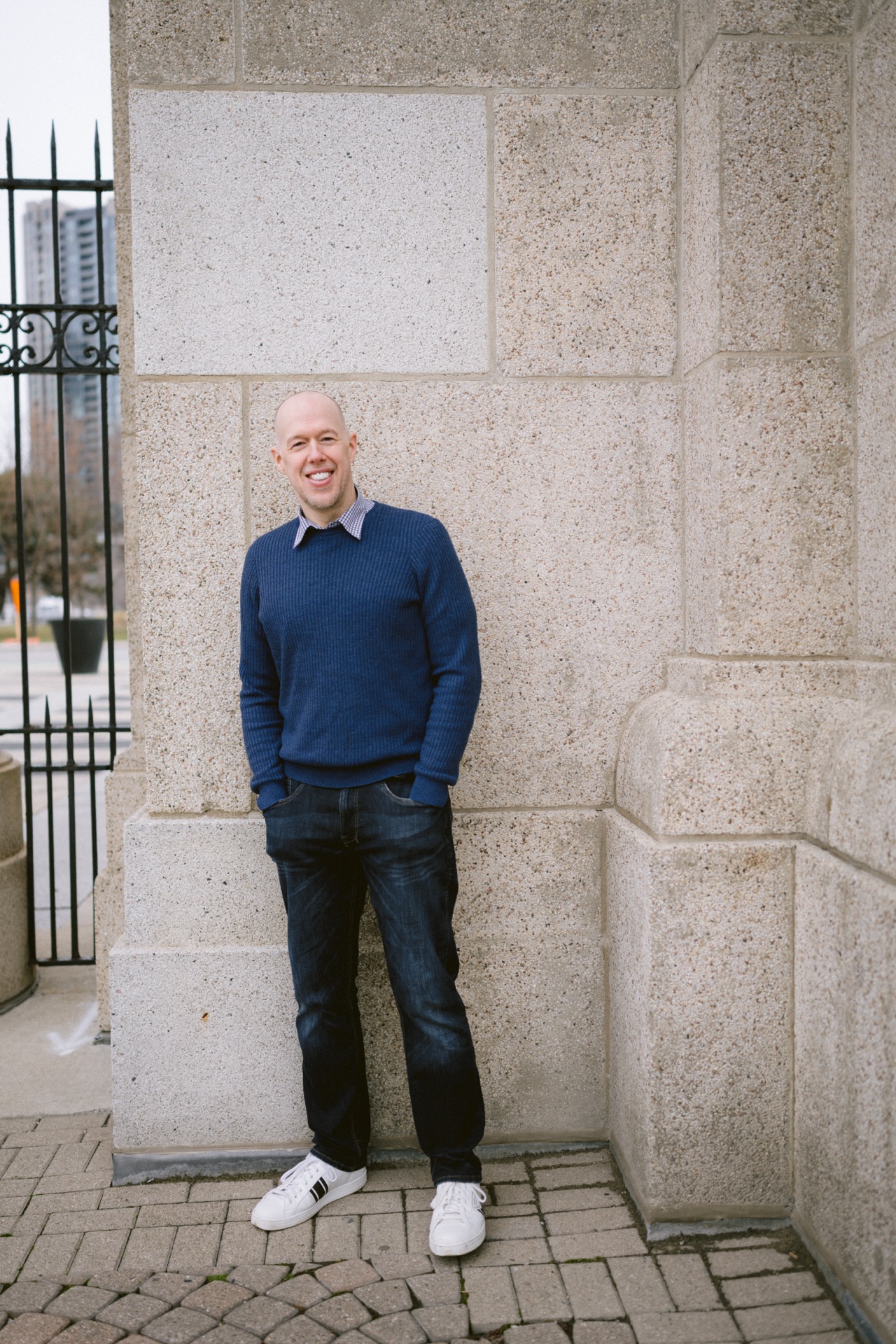 A smiling man in a blue sweater and jeans standing by a stone wall.