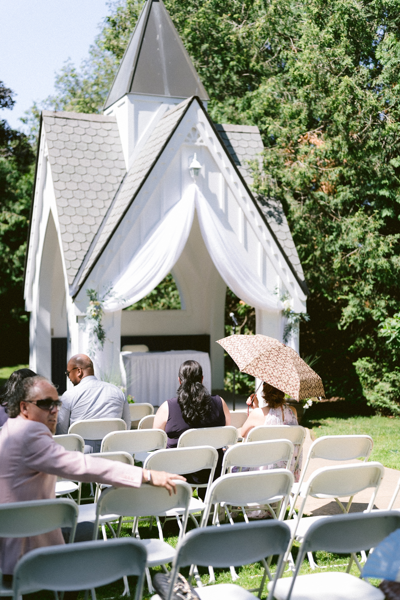 Guests seated at an outdoor wedding ceremony near a white gazebo on a sunny day.