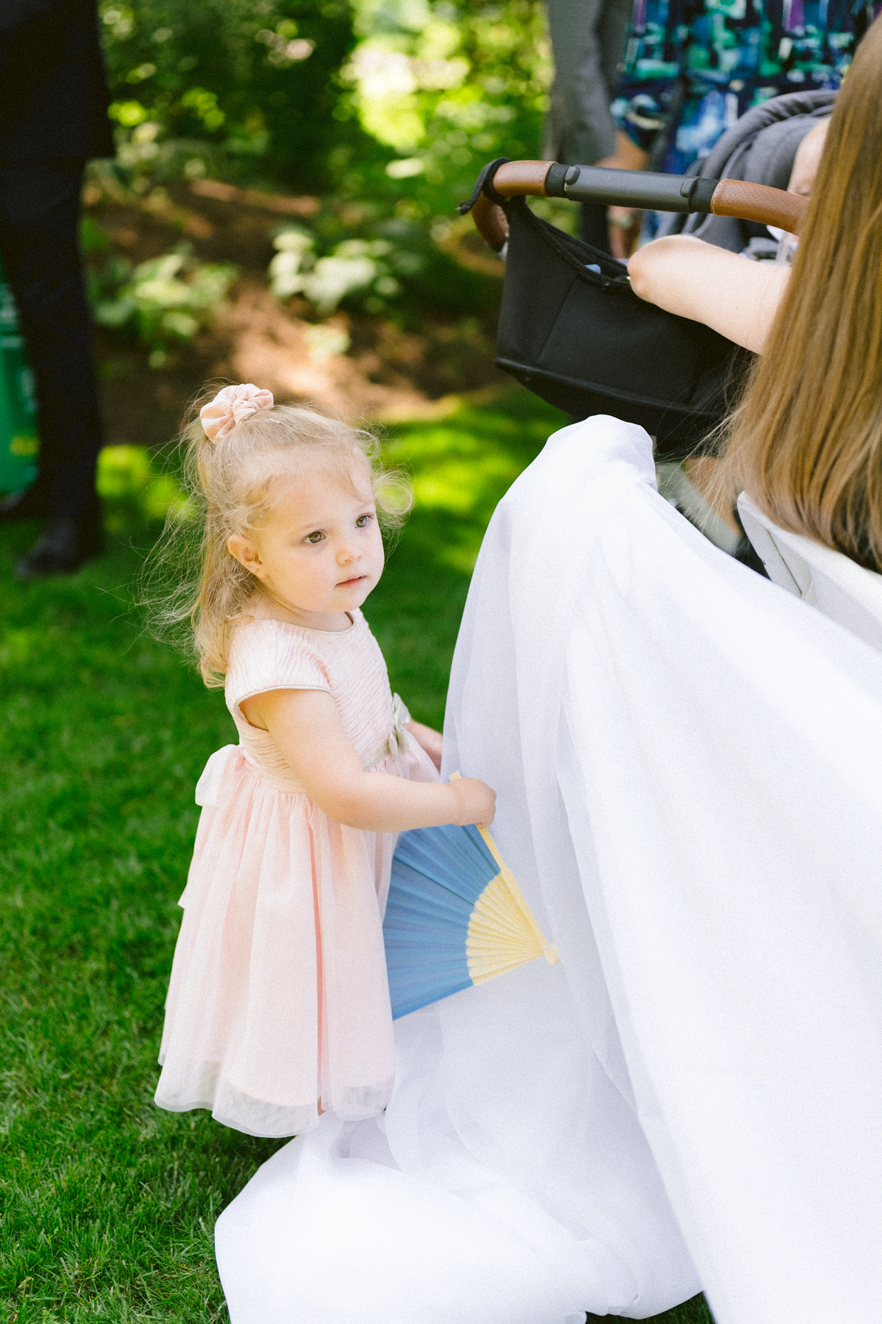 A young child in a pink dress curiously examines the white fabric of a dress at an outdoor gathering.