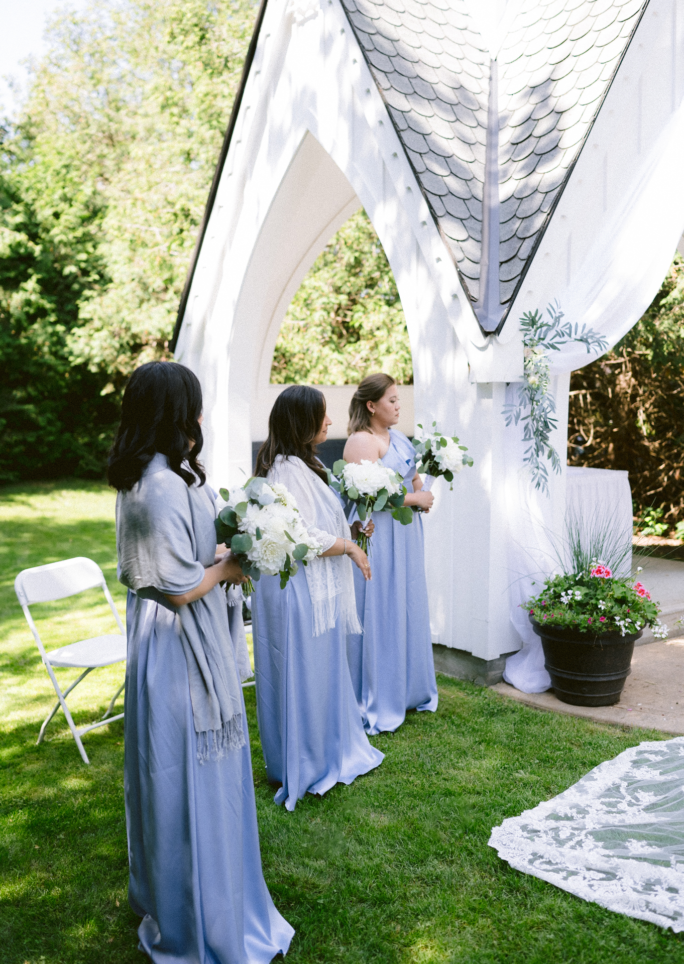 Three bridesmaids in blue dresses standing beside a white archway at an outdoor wedding ceremony.