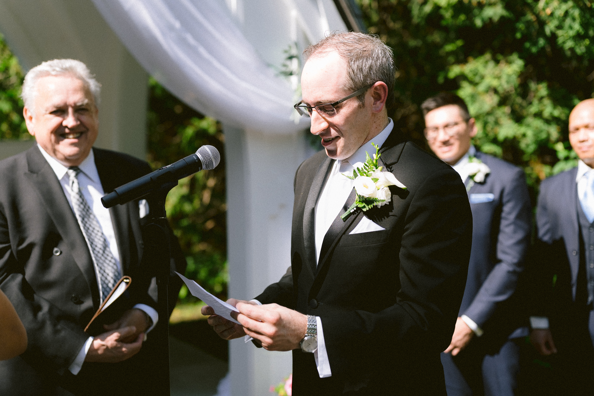 A man in a suit with a boutonniere reading from a paper at an outdoor wedding ceremony, with an officiant and groomsmen looking on.