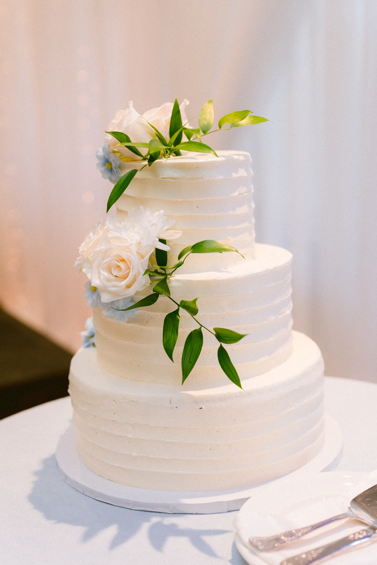Three-tiered wedding cake adorned with white flowers and green leaves, displayed on a table.