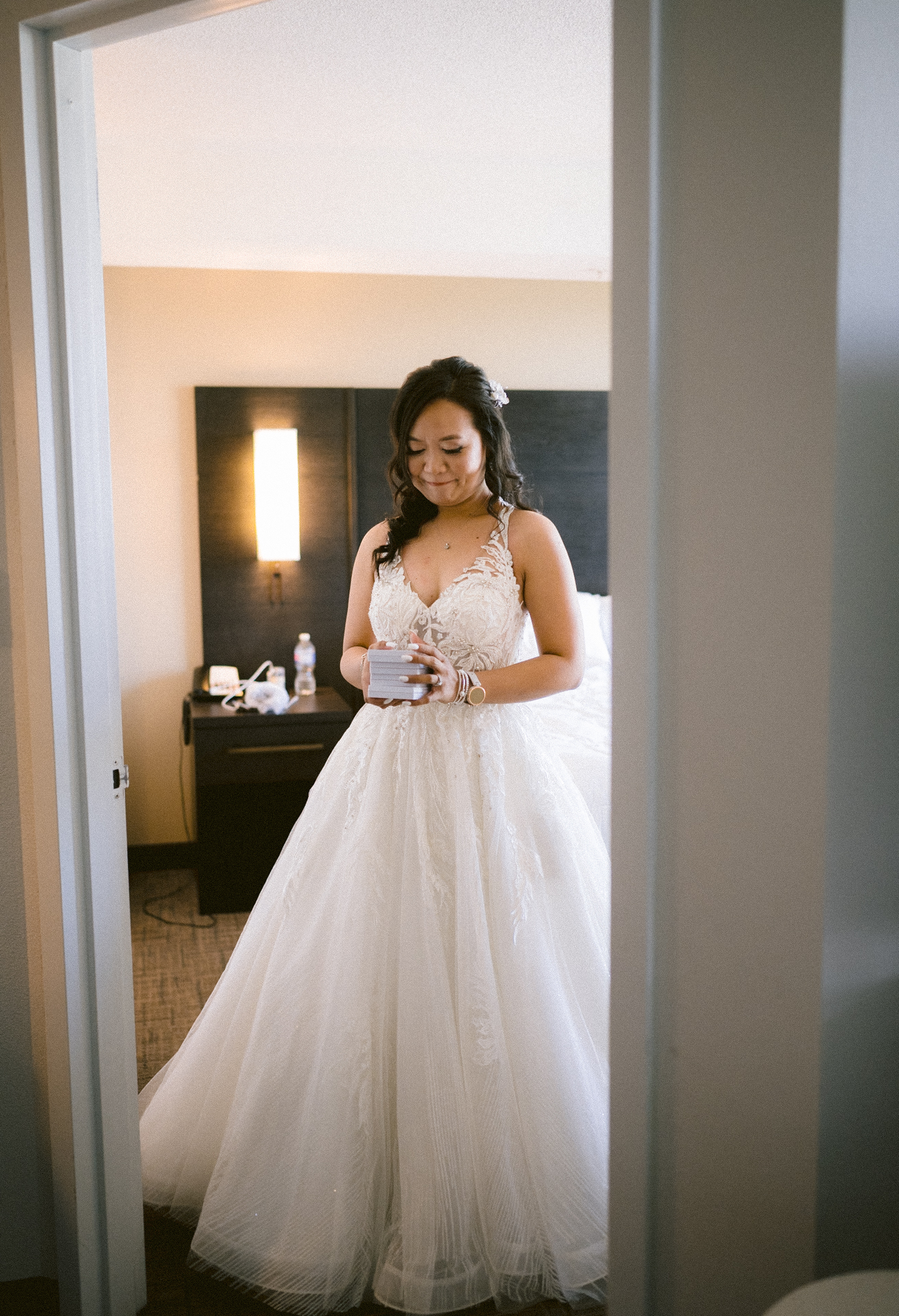 A bride in a white gown holding a cup, standing contemplatively in a hotel room.