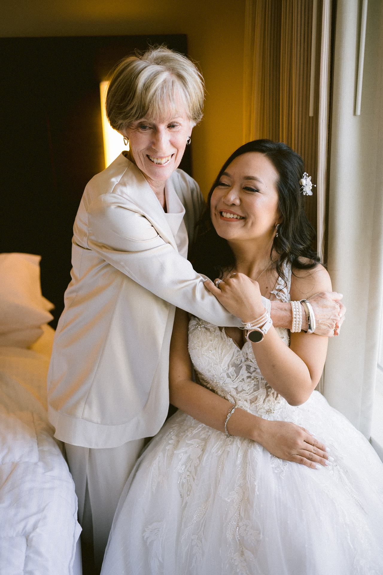 Two women embracing happily, one in a bridal gown and the other in formal attire.