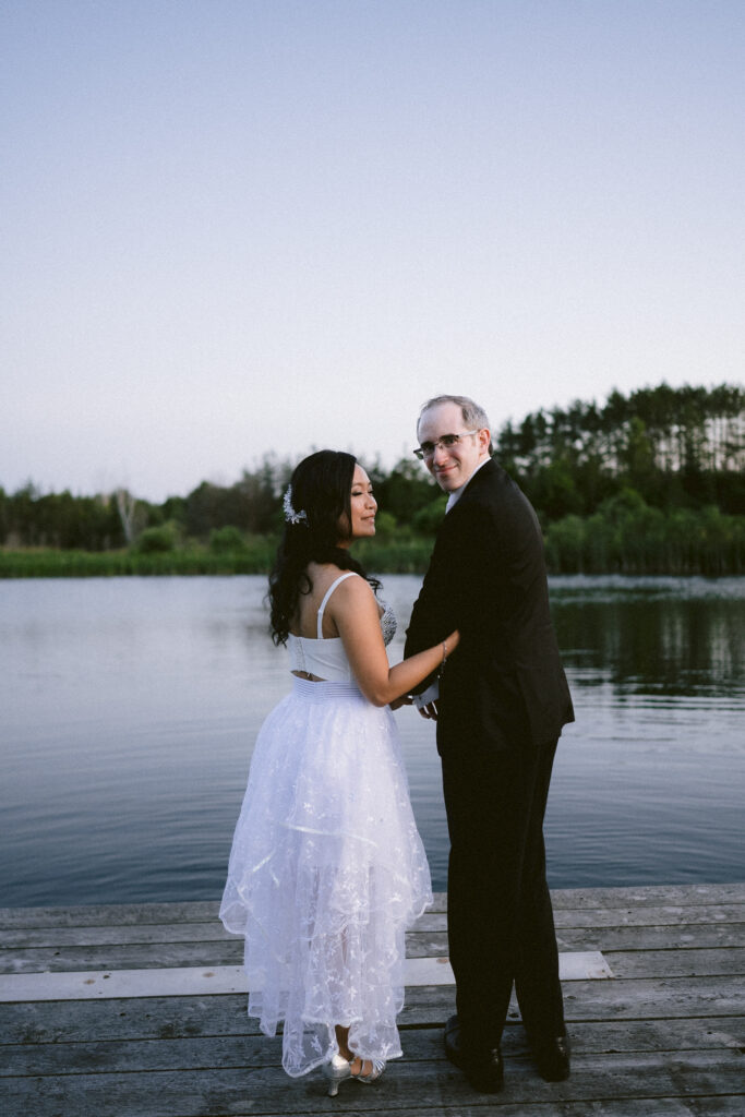 A couple in wedding attire sharing a moment on a wooden dock by a lake.