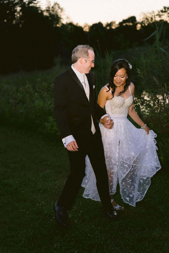 A bride and groom walking together and smiling in a twilight outdoor setting.
