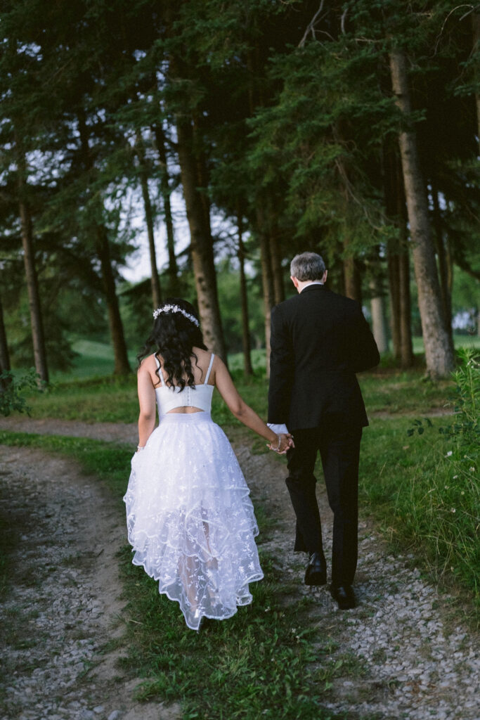 A couple holding hands, walking down a wooded path, with the person on the left wearing a white dress and the person on the right wearing a black suit.