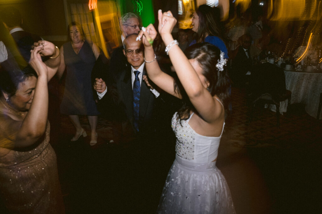 People dancing at an indoor celebration with a young female wearing a tiara leading the dance.