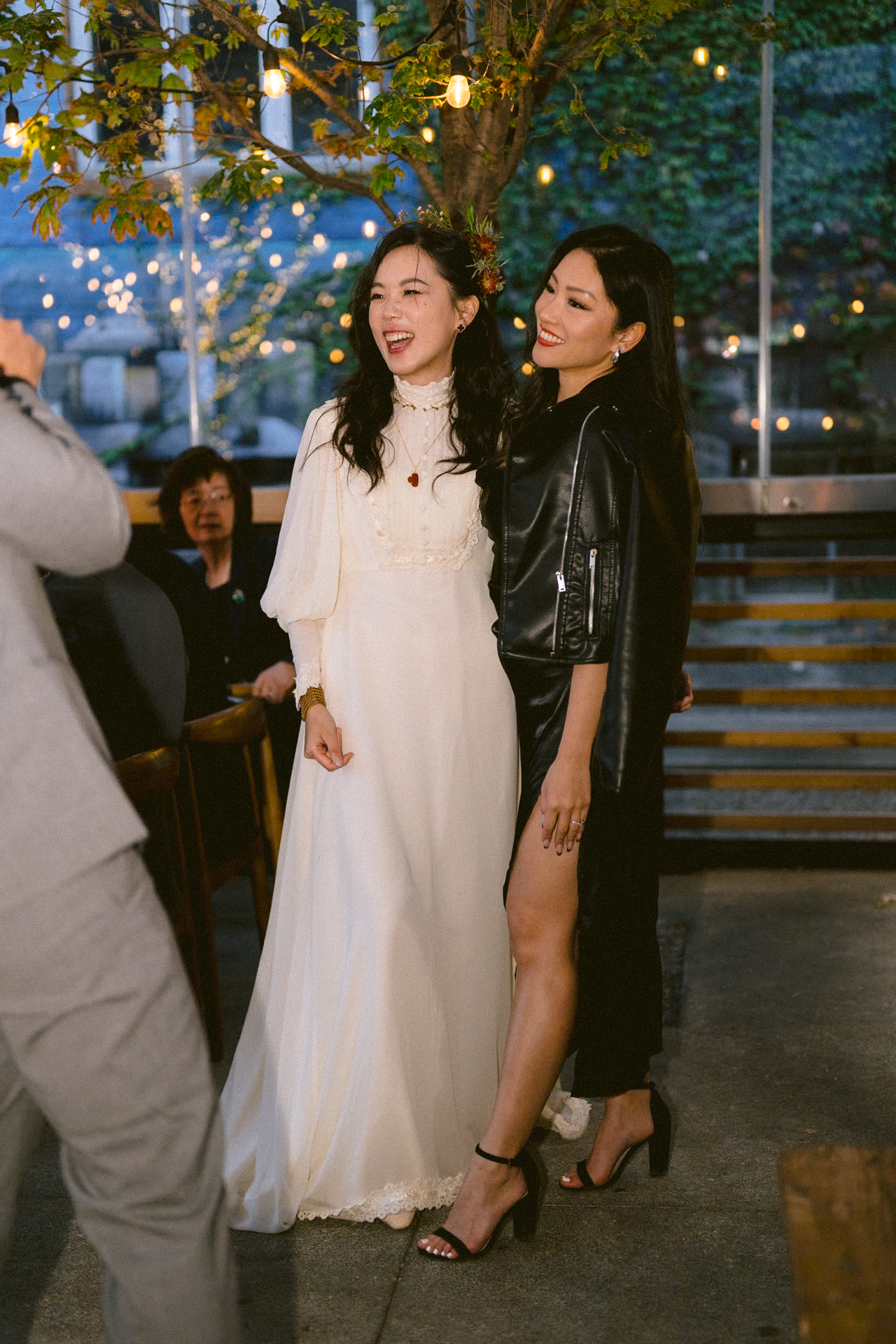 Two women posing for a photograph at an evening event, one in a white dress and the other in black attire, with string lights in the background.