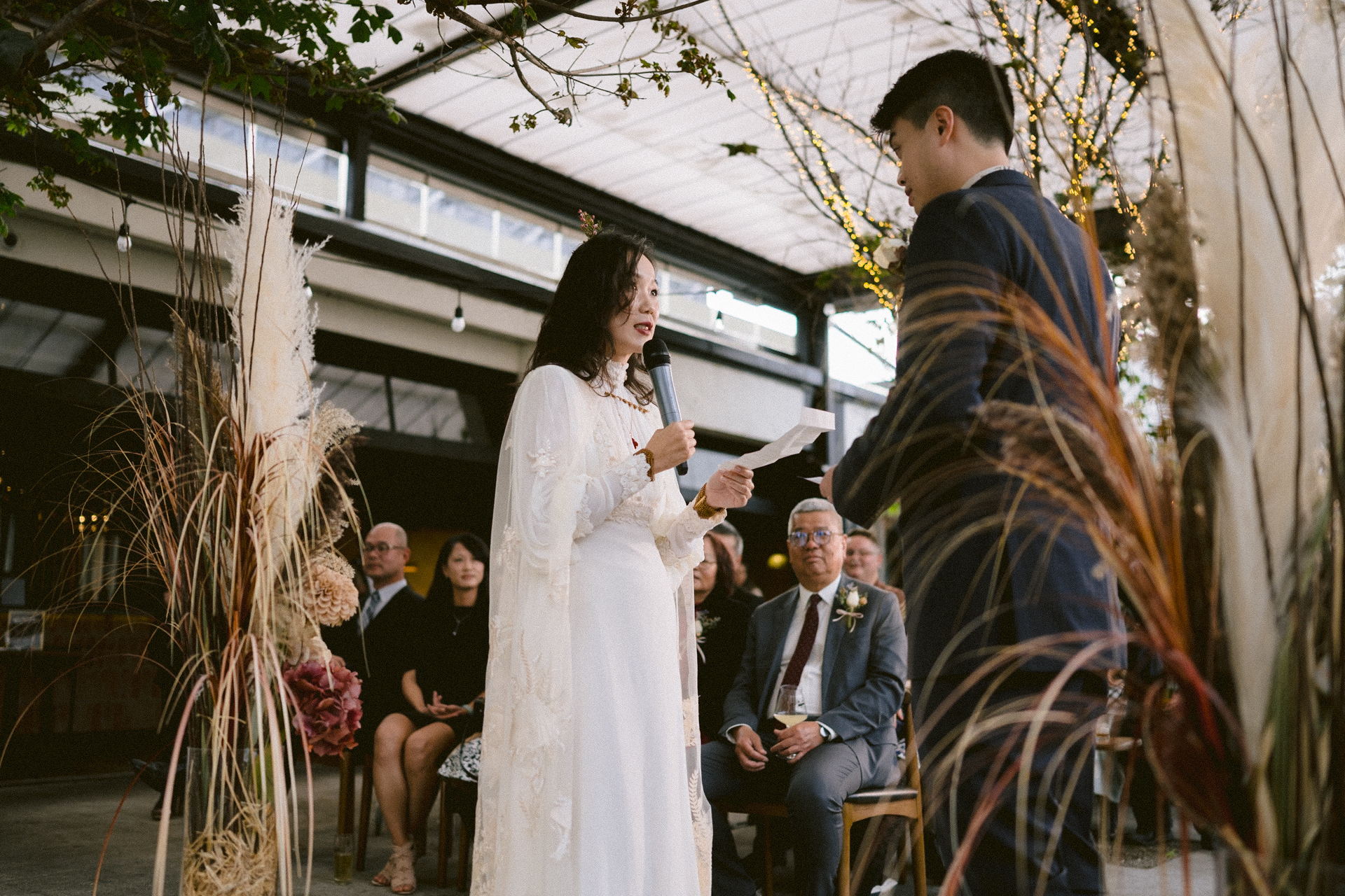 A bride reads her vows to the groom during an indoor wedding ceremony surrounded by guests and decorative foliage.