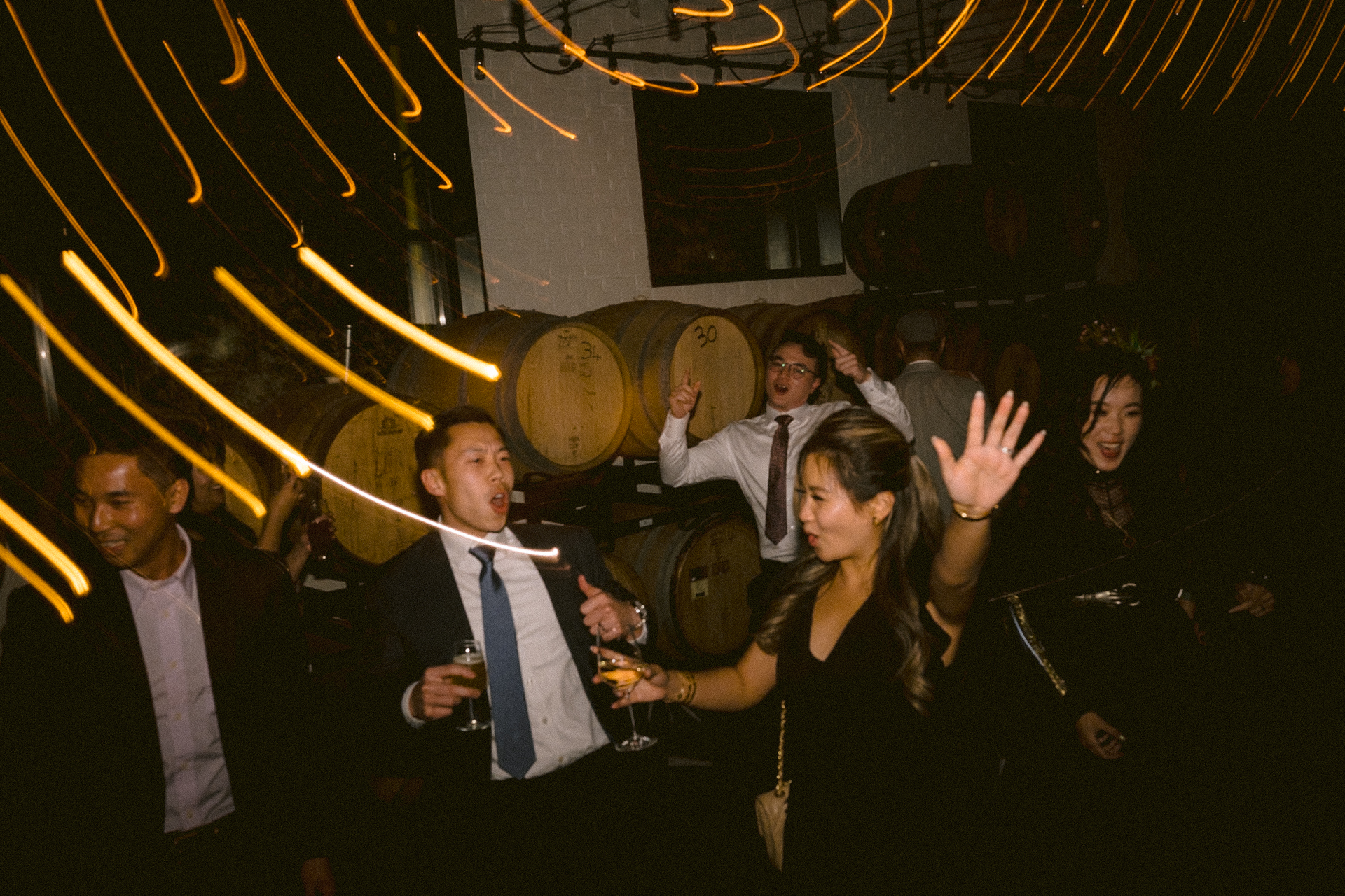 A group of people celebrating and dancing in a dimly lit room with decorative lights and barrels in the background.