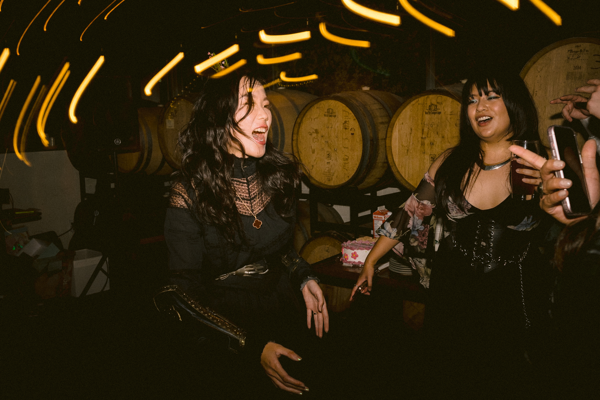 Bride and her friends laughing and enjoying themselves at a social event with wooden barrels in the background and ambient lighting.