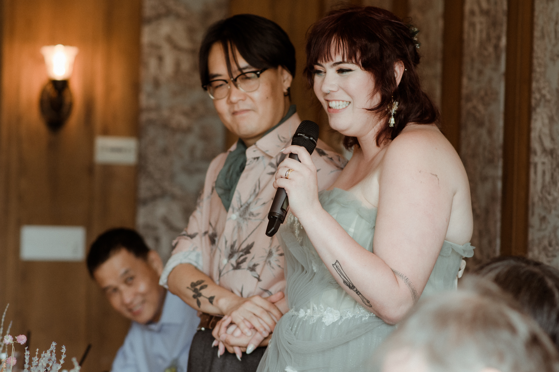 A woman holding a microphone gives a speech beside a man at a wedding reception.