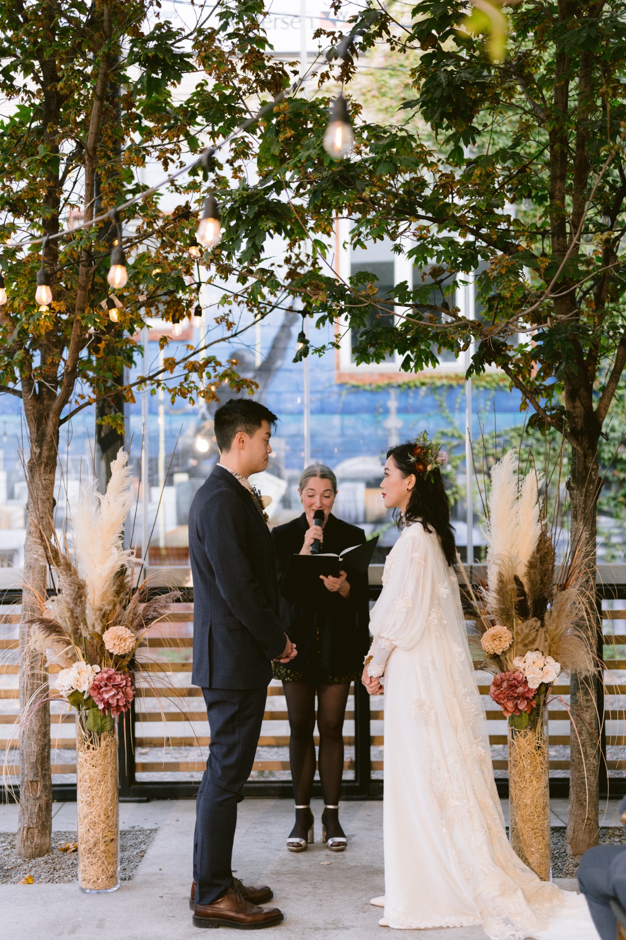 A couple exchanges wedding vows outdoors with an officiant presiding over the ceremony, surrounded by greenery and floral decorations