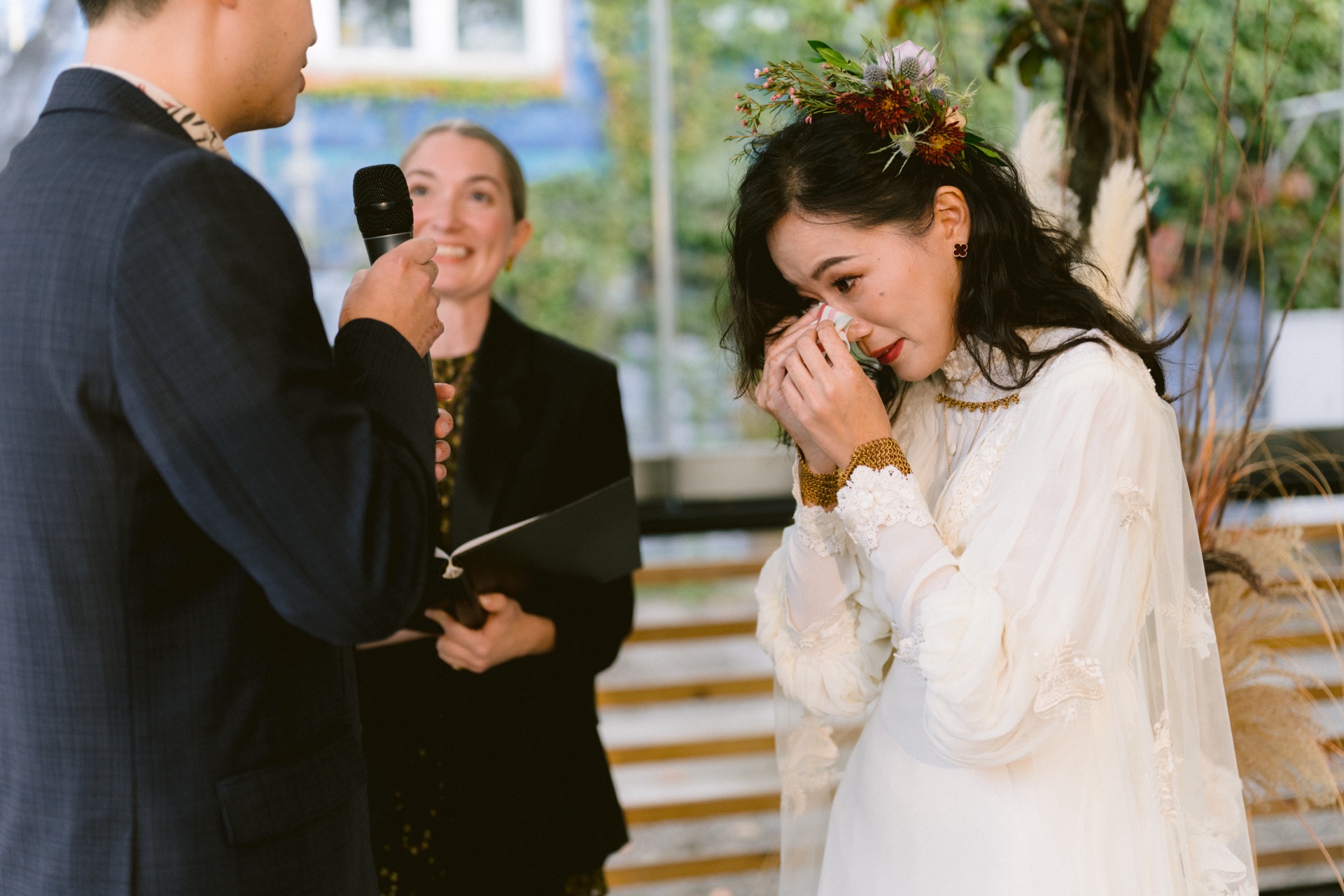 A bride wipes away tears during a wedding ceremony as the groom speaks and an officiant looks on