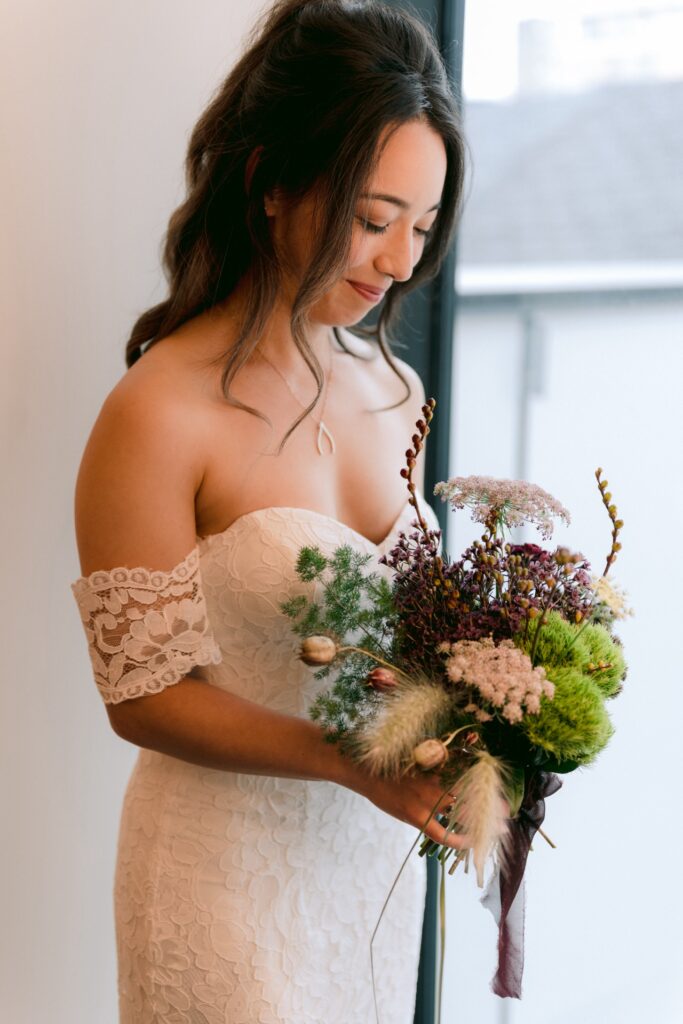 A bride in an off-shoulder lace dress holds a bouquet of wildflowers while gazing downwards.