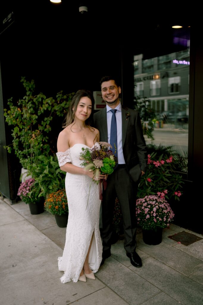A couple in wedding attire smiling together with a bouquet, standing next to potted flowers.