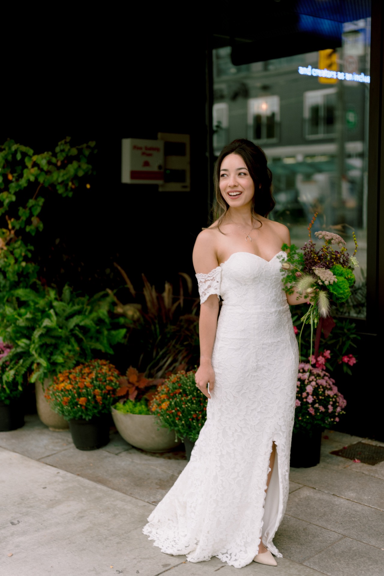 A bride in a white off-the-shoulder gown smiling and holding a bouquet on a city sidewalk.