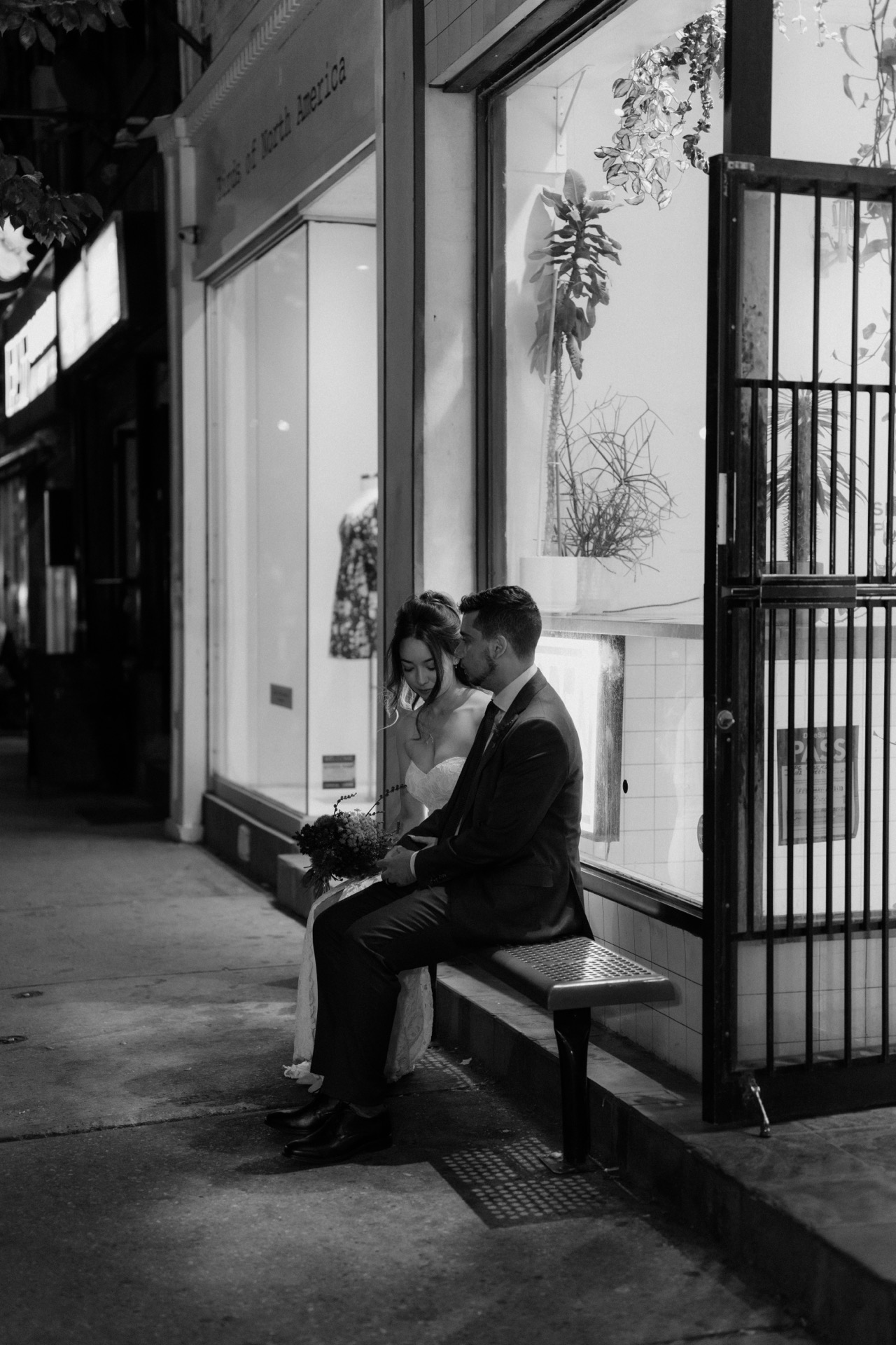 A couple in wedding attire sitting together on a bench at night outside a storefront.