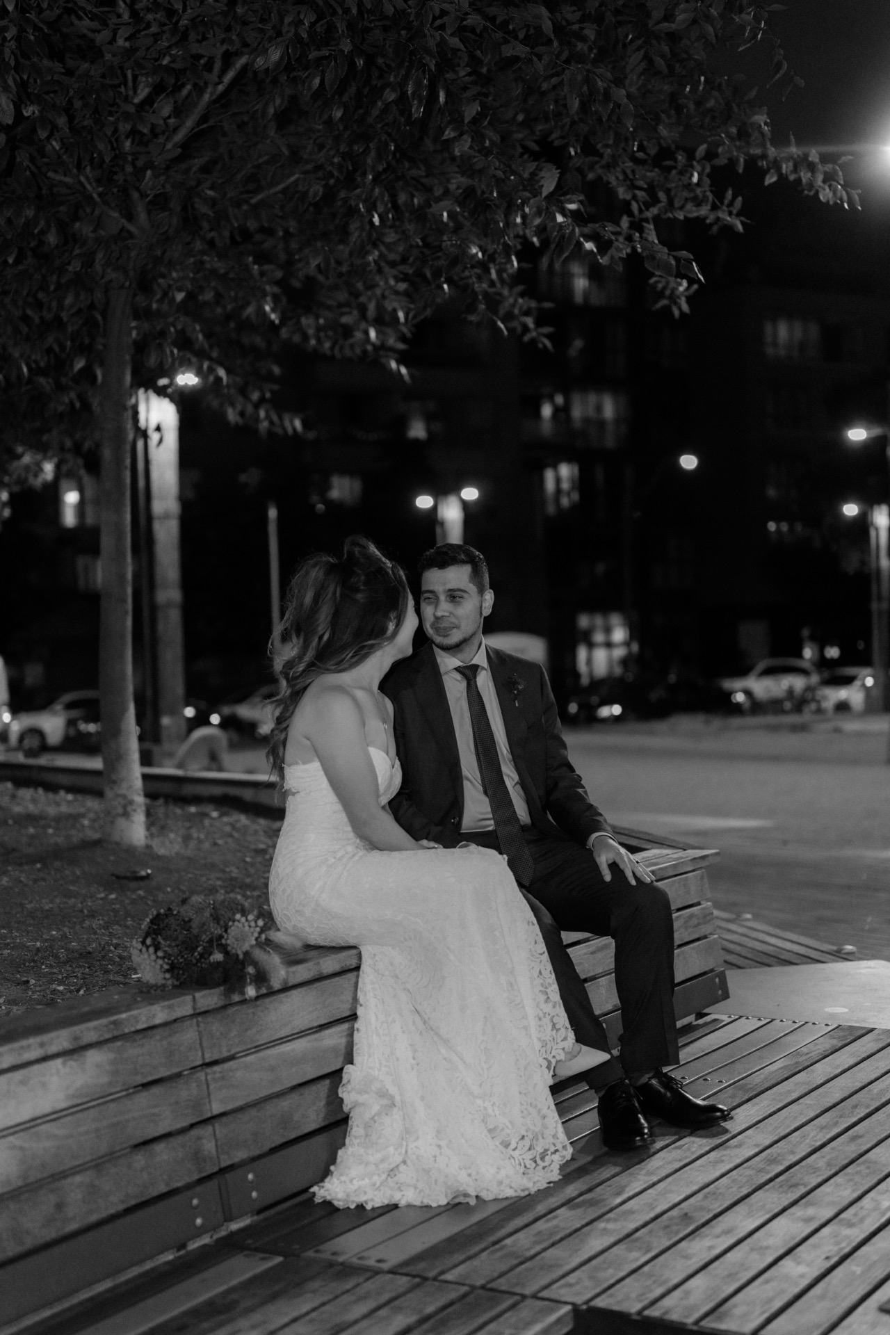 A couple in wedding attire sitting together on a bench at night