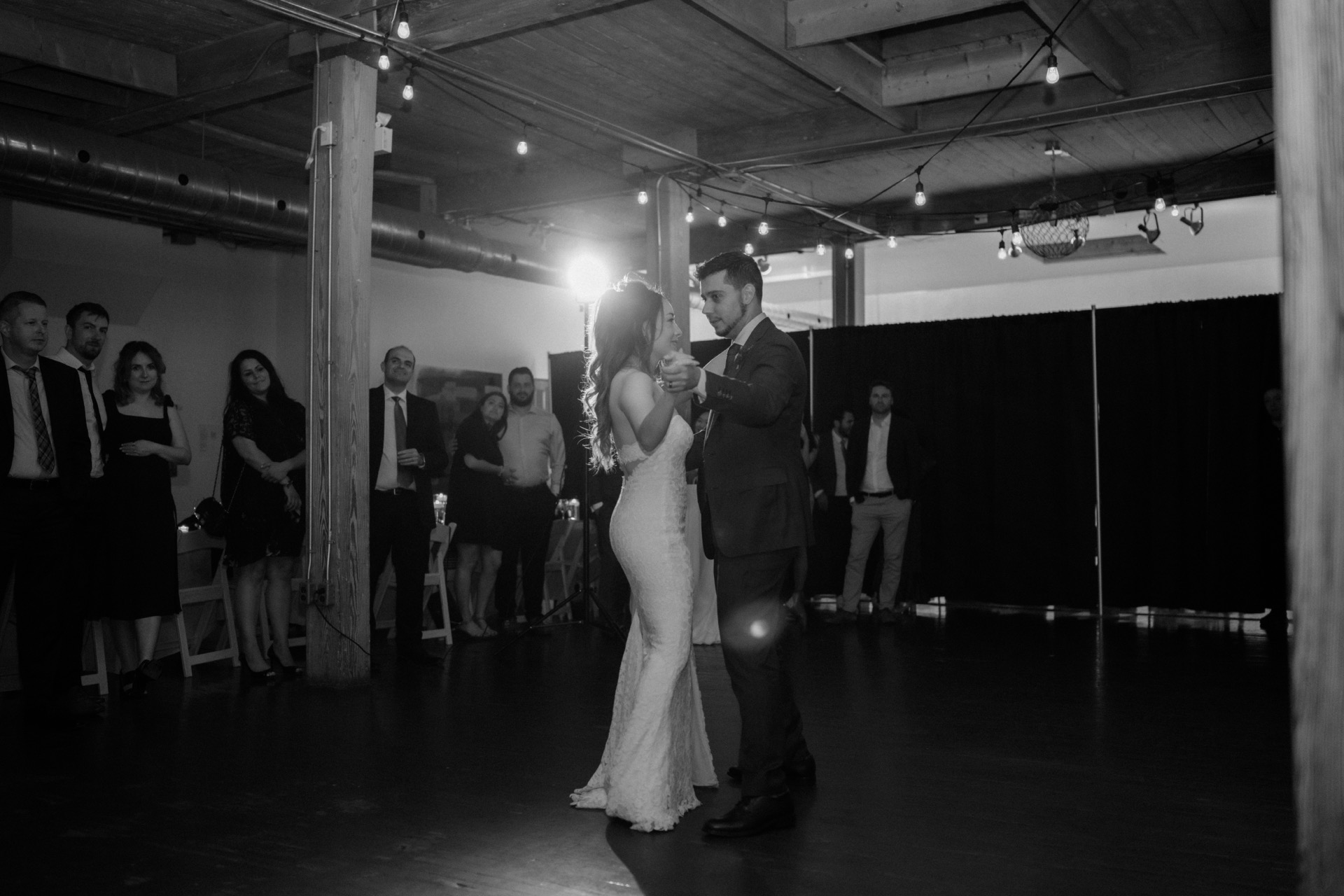 A couple shares a dance in a dimly lit room as onlookers surround them.