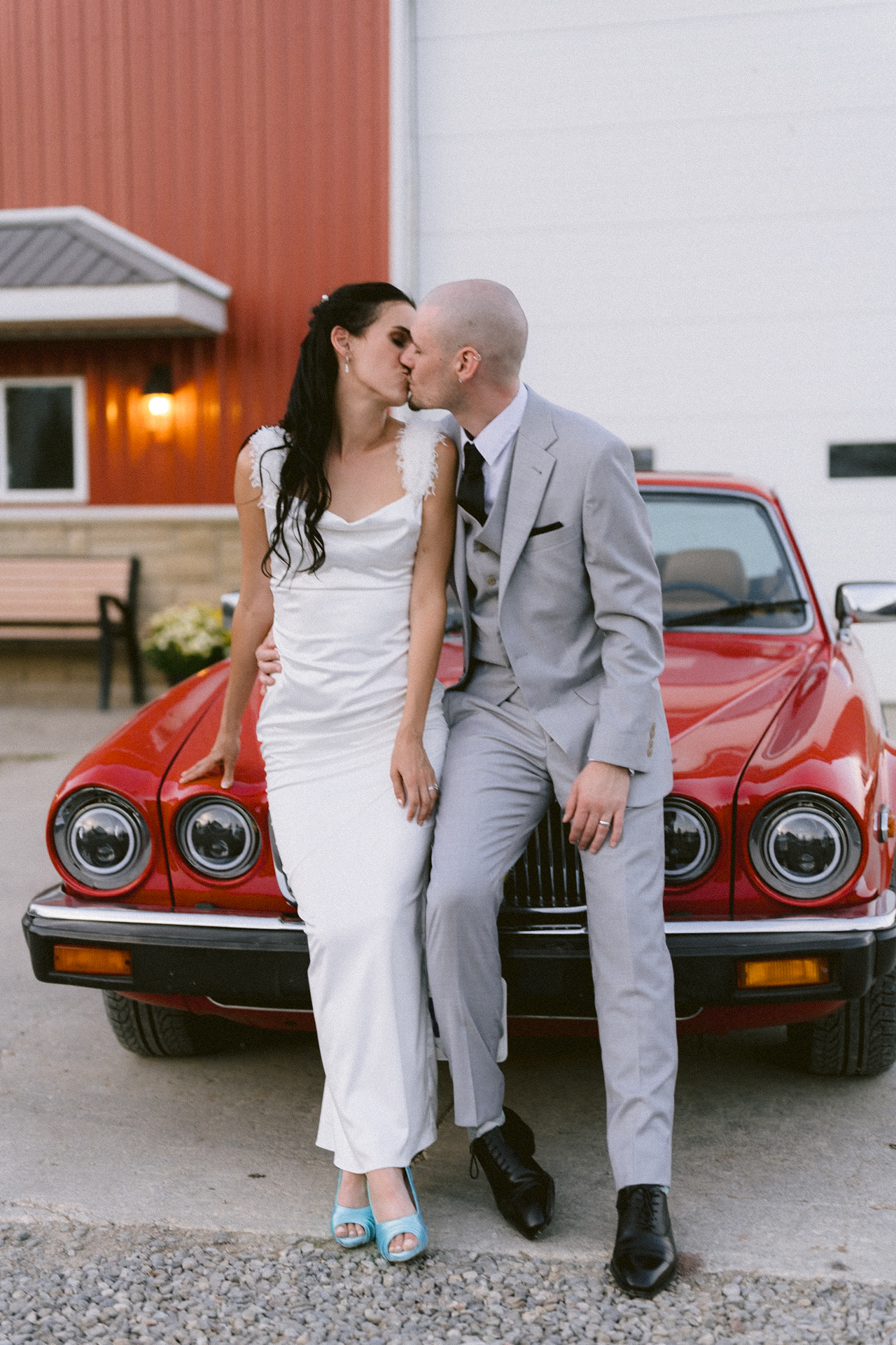 Newlyweds sharing a kiss in front of a classic red car.