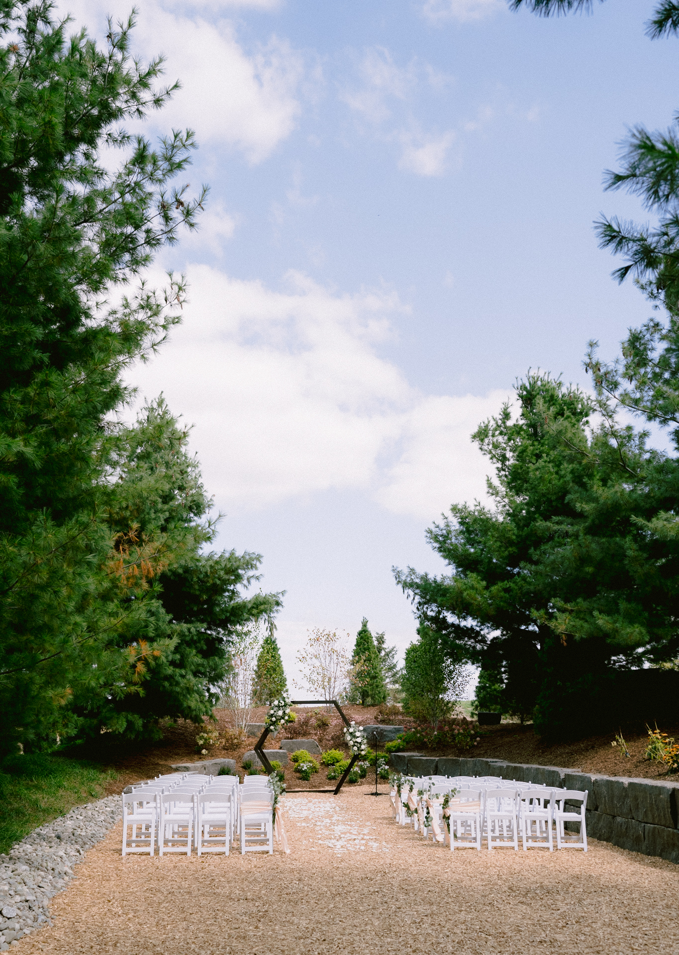 Outdoor wedding ceremony setup with white chairs arranged on a gravel path, surrounded by greenery under a clear sky at Cambium Farms.
