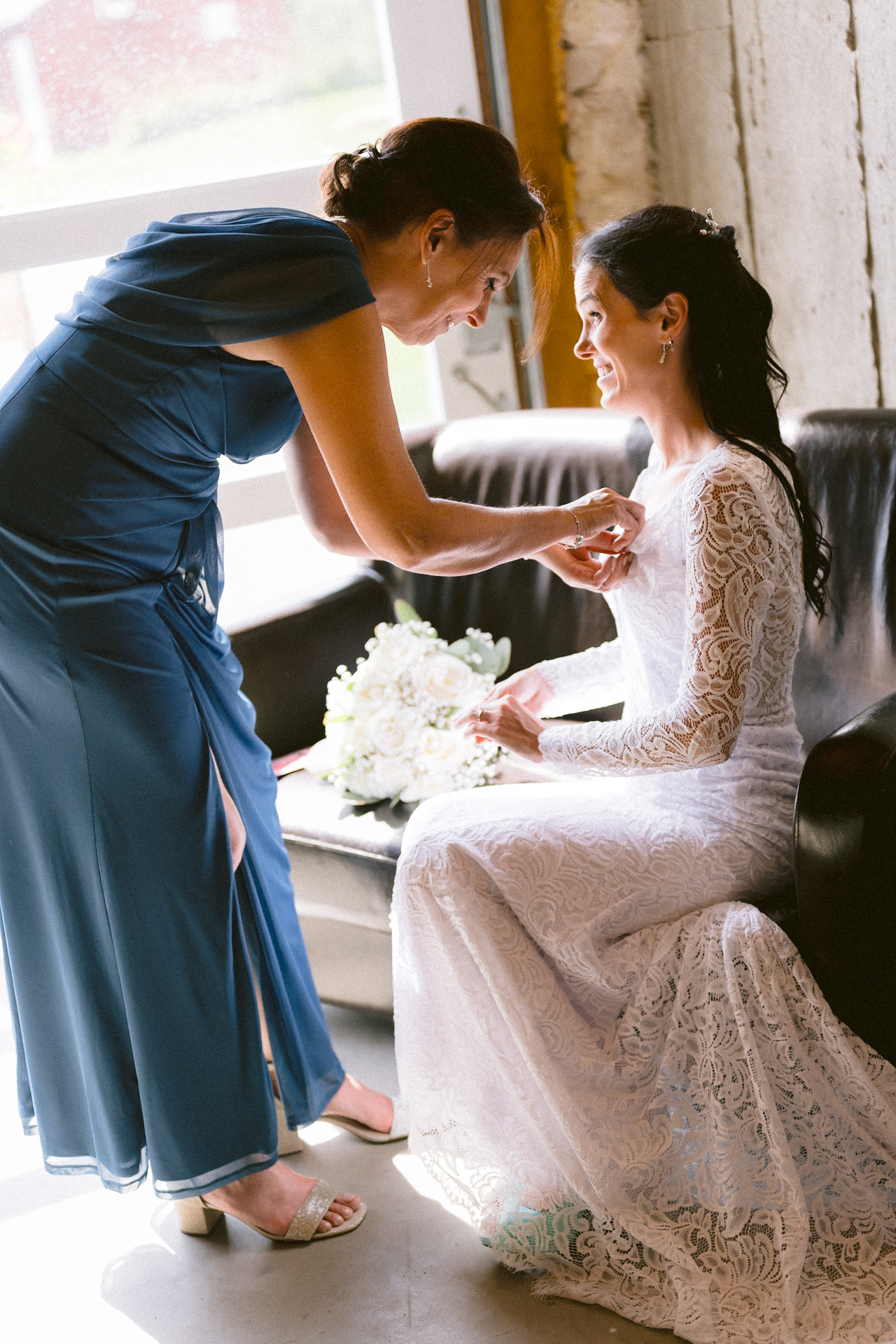 A woman in a blue dress adjusts the veil of a bride seated in a chair, both are indoors by a large window, with a bouquet on the table next to them.