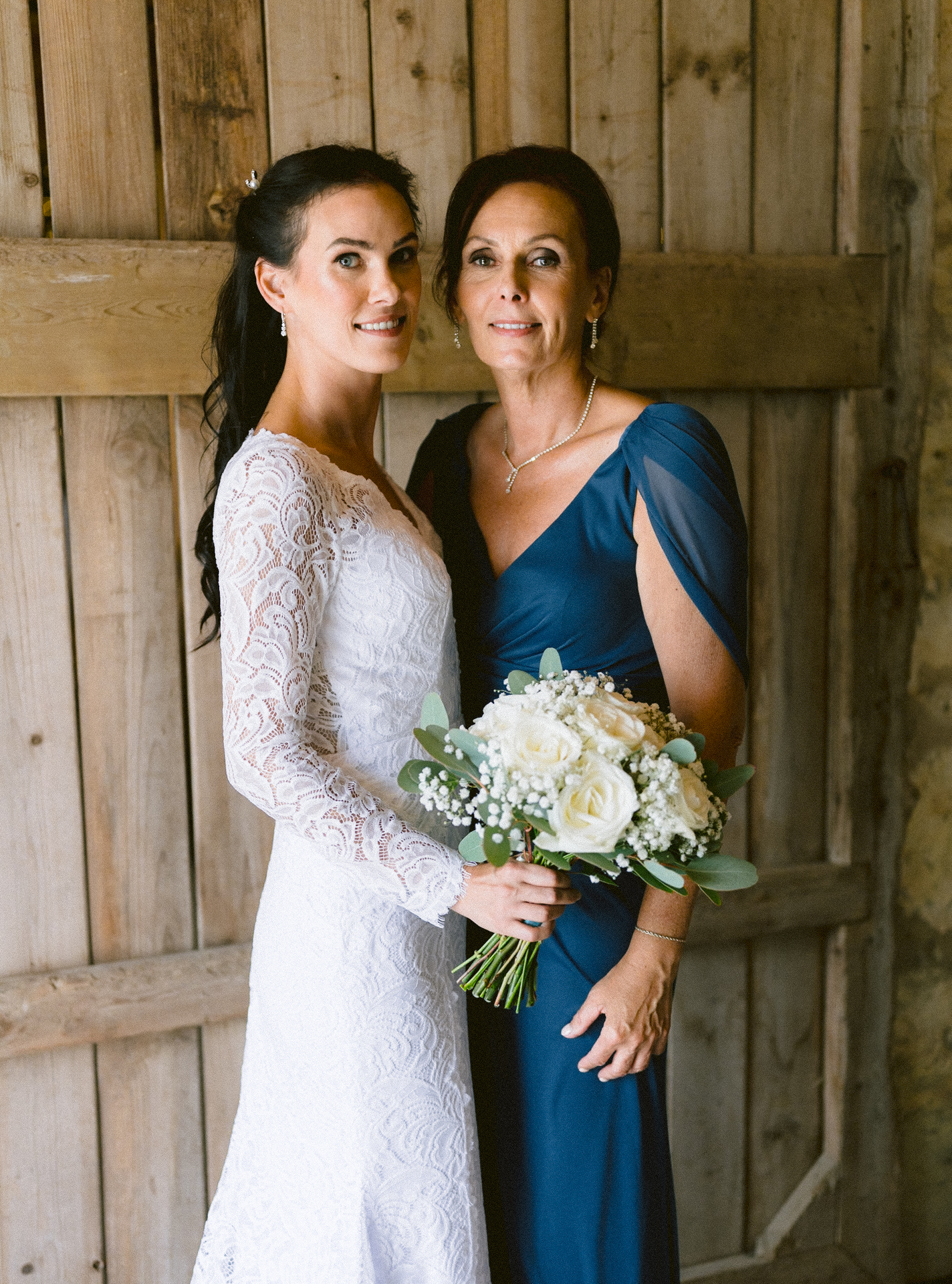 A bride in a white lace dress holding a bouquet stands next to her mother in a blue gown against a wooden backdrop.