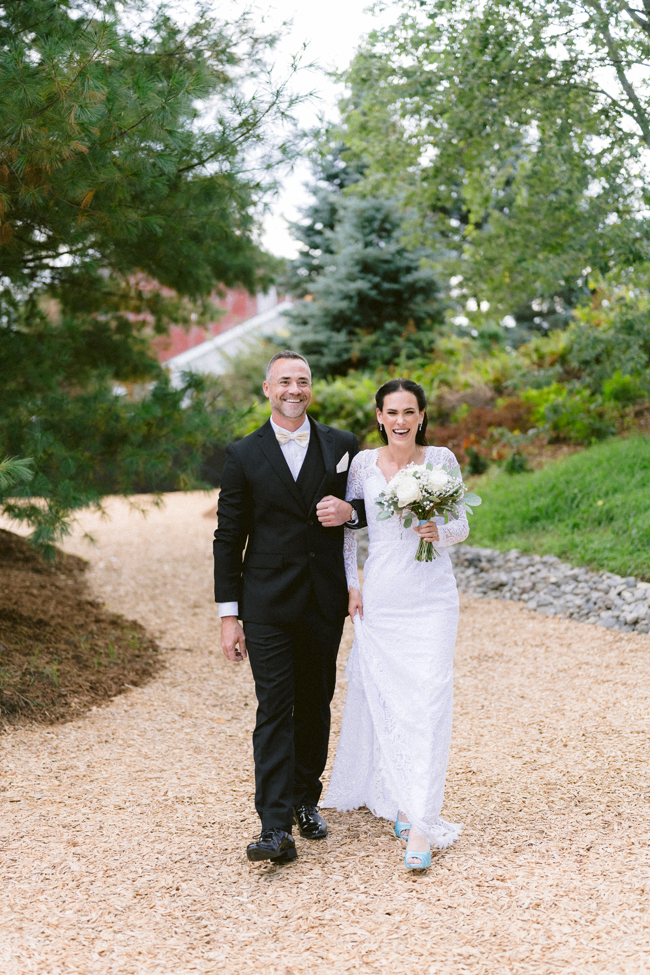 A smiling bride in wedding attire holding her brother walking down the aisle on a gravel path at Cambium Farms.