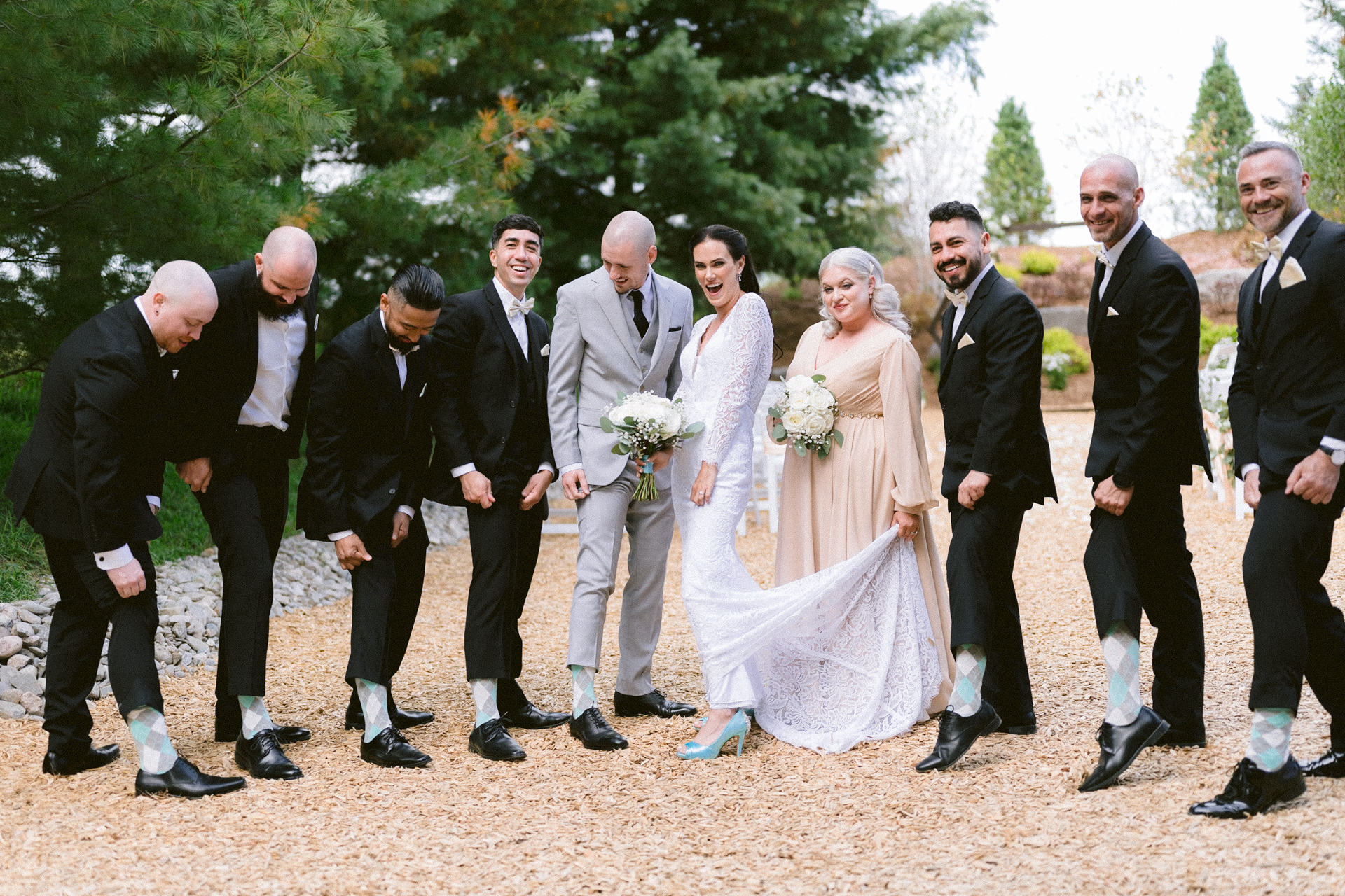 A wedding party posing whimsically with the bride and groom in the center, surrounded by groomsmen and a bridesmaid showcasing their socks and shoes.