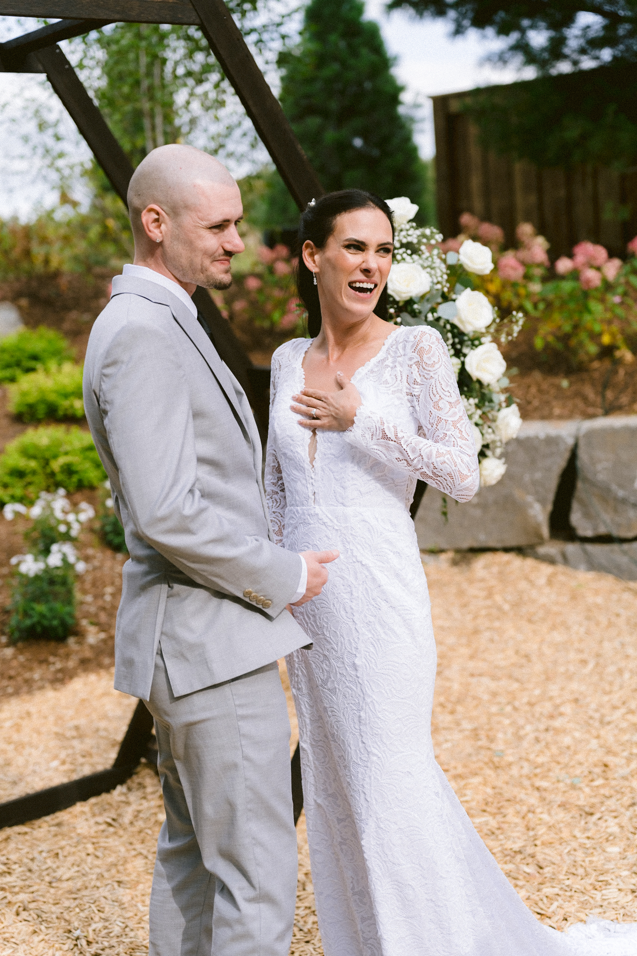 A smiling couple dressed in wedding attire, with the individual in a white dress and the other in a gray suit, stand together outdoors.